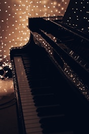 A close-up of a piano keyboard with a shiny black surface reflects strings of warm white fairy lights in the background, creating a serene and cozy atmosphere.