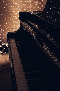 A close-up of a piano keyboard with a shiny black surface reflects strings of warm white fairy lights in the background, creating a serene and cozy atmosphere.