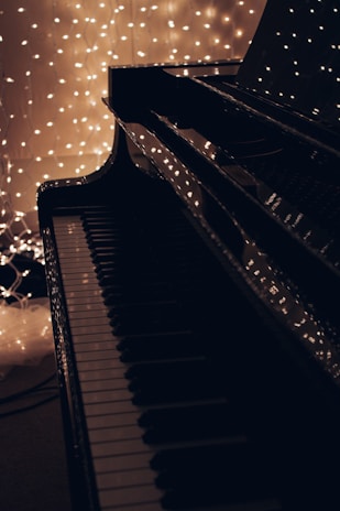 A close-up of a piano keyboard with a shiny black surface reflects strings of warm white fairy lights in the background, creating a serene and cozy atmosphere.