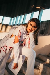 A person with long hair in a relaxed pose, leaning on one hand, dressed in a stylish outfit with a mix of white and floral patterns. The background includes a series of large windows with sunlight streaming in, casting strong shadows.