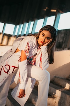 A person with long hair in a relaxed pose, leaning on one hand, dressed in a stylish outfit with a mix of white and floral patterns. The background includes a series of large windows with sunlight streaming in, casting strong shadows.