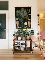 A cozy interior setting features a wooden stand with several potted plants on top, displaying a variety of greenery. Below, a grey cat comfortably lies on the lower shelf beside vintage cameras. A decorative wall hanging depicting botanical illustrations is prominently displayed above the stand. A wicker chair with a soft cushion sits nearby, contributing to the inviting atmosphere.
