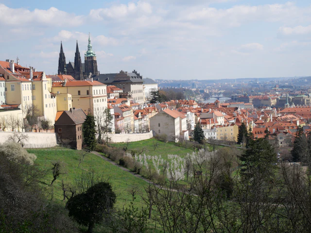 A view across red-roofed Hradcany rooftops toward St. Vitus Cathedral's spires and Prague Castle on a bright day.