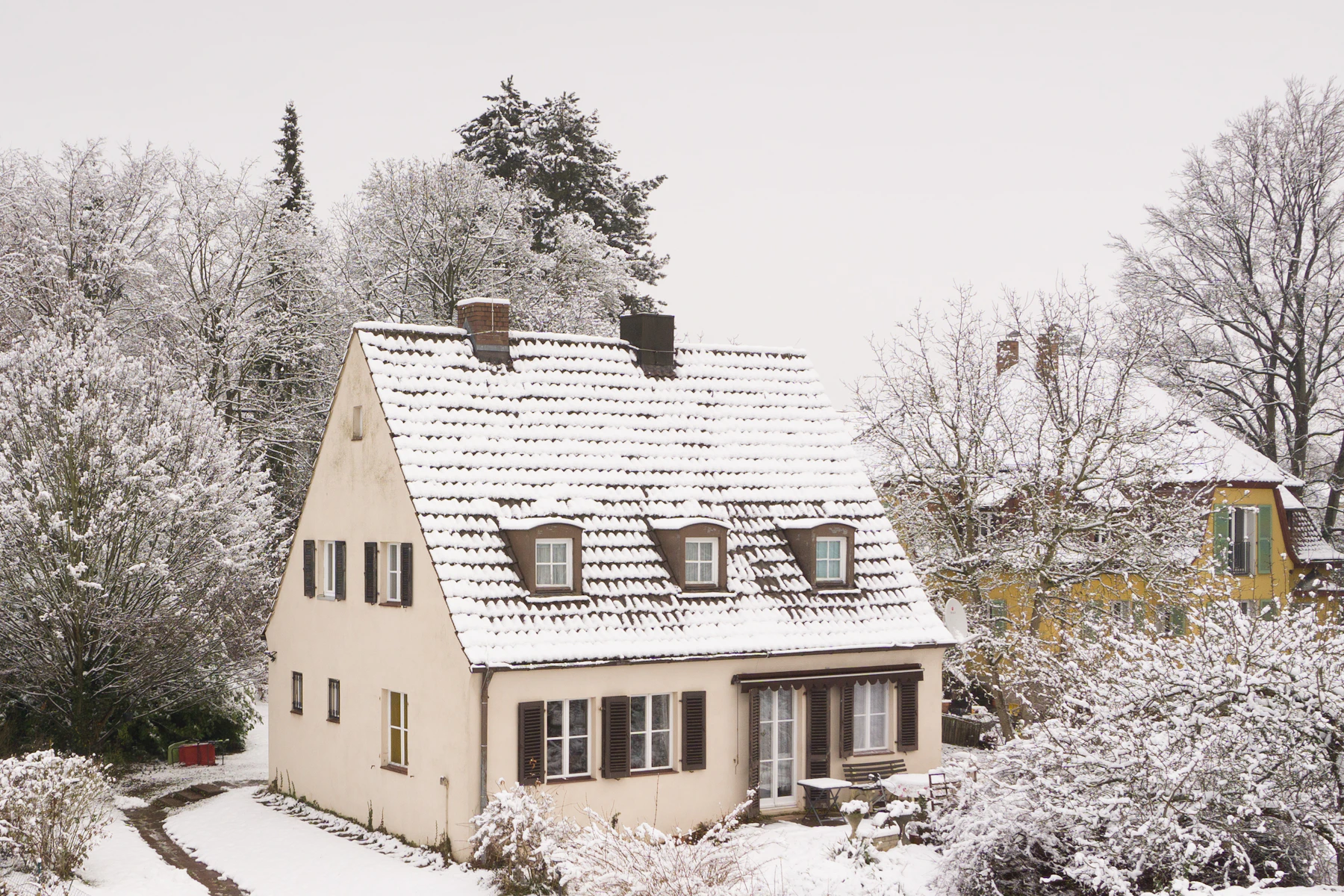 A cream cottage with dormer windows and a snow-covered roof among frosted trees