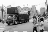 A large truck moves along a city street surrounded by tall buildings. Several pedestrians are visible in the foreground, some holding papers. The truck has a promotional banner on its side. The scene is urban and lively, with vehicles and people sharing the space.