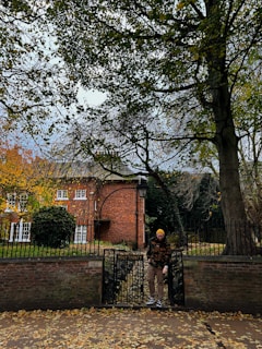A person is standing in front of a black wrought iron gate with the words 'Holy Trinity' mounted on it. Behind the gate, there is a brick building surrounded by trees with autumn foliage. The ground is covered with fallen leaves, and a large tree frames the scene on the right.