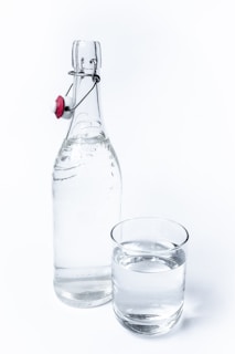 A clear glass bottle with a flip-top cap sits next to a glass of water. The bottle has a metallic clasp with a red and white ceramic stopper. The glass is filled with water to near the brim, and both items are set against a plain white background.