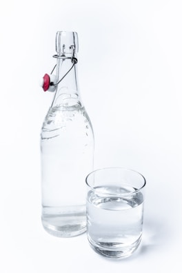 A clear glass bottle with a flip-top cap sits next to a glass of water. The bottle has a metallic clasp with a red and white ceramic stopper. The glass is filled with water to near the brim, and both items are set against a plain white background.