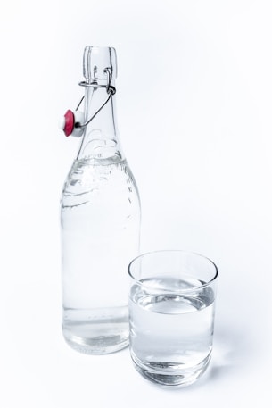 A clear glass bottle with a flip-top cap sits next to a glass of water. The bottle has a metallic clasp with a red and white ceramic stopper. The glass is filled with water to near the brim, and both items are set against a plain white background.