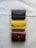 A set of three leather wallets arranged vertically on a light fabric surface. The wallets are of different colors: dark brown, mustard yellow, and reddish-brown. Each wallet has a flap secured with a metal button.