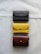 A set of three leather wallets arranged vertically on a light fabric surface. The wallets are of different colors: dark brown, mustard yellow, and reddish-brown. Each wallet has a flap secured with a metal button.