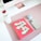 A neatly arranged desktop setup features a large monitor with a rose gold stand, a white water bottle with a red cap, a pink notebook titled 'You're Doing So Well', a pair of wireless earbuds, and a smartphone. The items are placed on a soft pink desk mat.