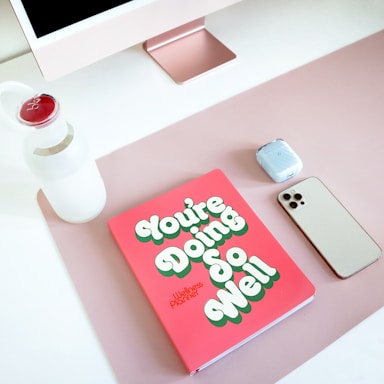 A neatly arranged desktop setup features a large monitor with a rose gold stand, a white water bottle with a red cap, a pink notebook titled 'You're Doing So Well', a pair of wireless earbuds, and a smartphone. The items are placed on a soft pink desk mat.