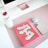 A neatly arranged desktop setup features a large monitor with a rose gold stand, a white water bottle with a red cap, a pink notebook titled 'You're Doing So Well', a pair of wireless earbuds, and a smartphone. The items are placed on a soft pink desk mat.