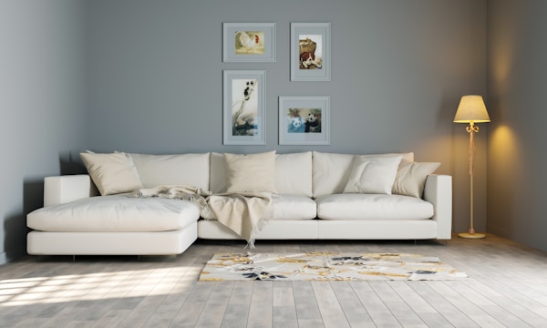 A modern living room features a white sectional sofa adorned with several throw pillows. Above the sofa, four framed art pieces are arranged neatly on the gray wall. To the right, a floor lamp with a warm glowing lampshade stands on the wooden floor. A patterned rug lies on the floor in front of the sofa, adding a touch of color.
