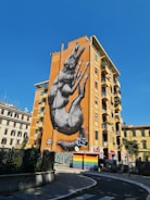 A large mural of a howling wolf is painted on the side of an orange-colored building. The artwork is highly detailed and dominates the building's facade. Surrounding structures include a mix of residential and commercial buildings, some with graffiti. A bright blue sky and clear weather enhance the visibility of the mural.