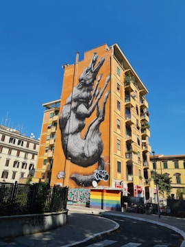 A large mural of a howling wolf is painted on the side of an orange-colored building. The artwork is highly detailed and dominates the building's facade. Surrounding structures include a mix of residential and commercial buildings, some with graffiti. A bright blue sky and clear weather enhance the visibility of the mural.