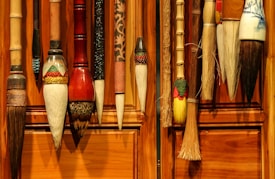 A collection of traditional calligraphy brushes hangs against a wooden wall. These brushes come in various sizes and designs, showcasing intricate craftsmanship with decorative elements on the handles.