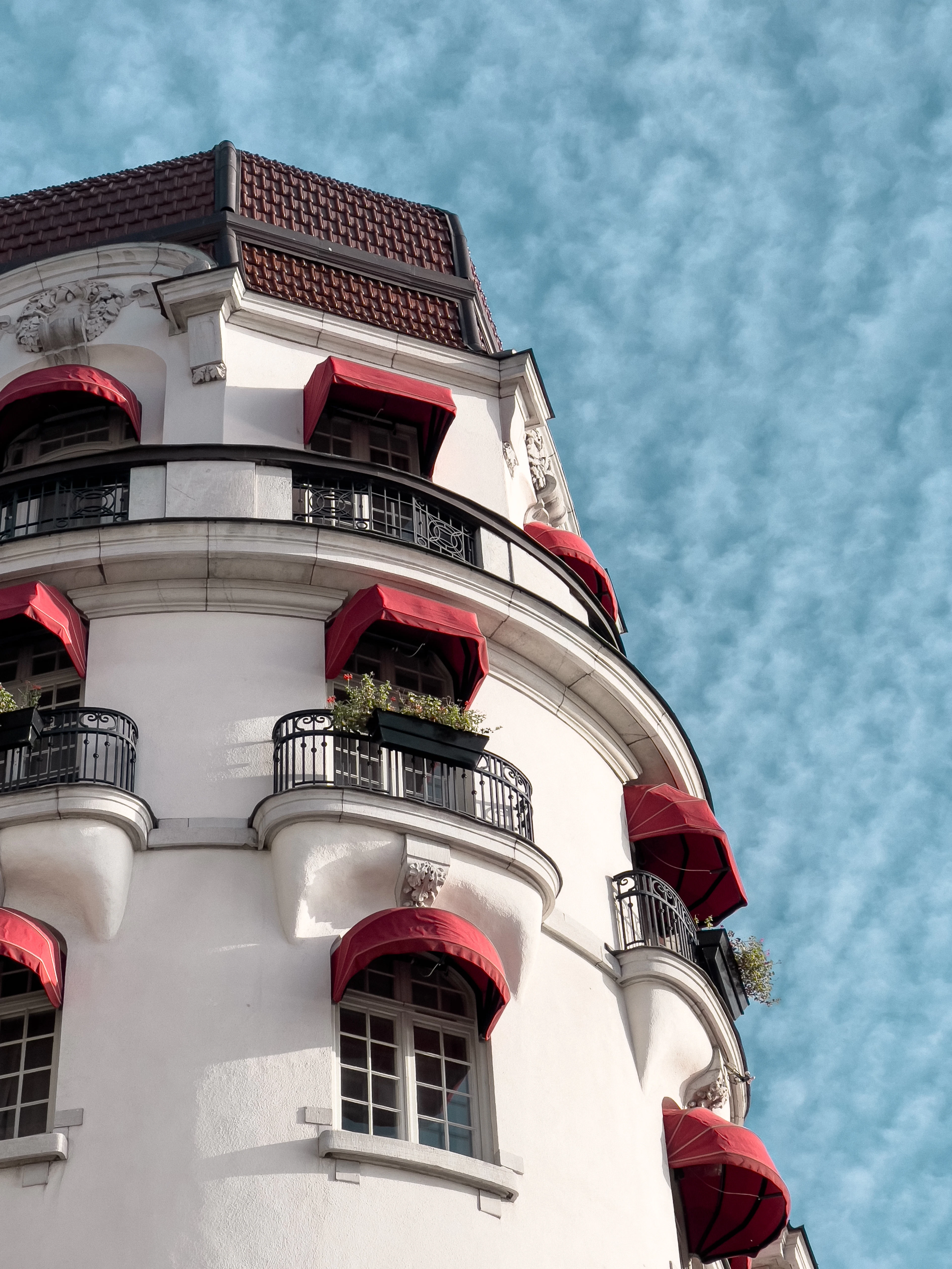 The curved white corner facade of a Stockholm hotel, its windows shaded by deep red awnings above wrought-iron balconies, set against a rippled mackerel sky.