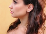 A profile of a person featuring long, dark hair and silver hoop earrings against a soft yellow background. The skin tone is smooth, and the hair slightly waves around the shoulders.
