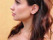 A profile of a person featuring long, dark hair and silver hoop earrings against a soft yellow background. The skin tone is smooth, and the hair slightly waves around the shoulders.