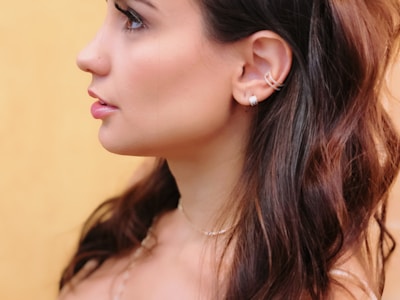 A profile of a person featuring long, dark hair and silver hoop earrings against a soft yellow background. The skin tone is smooth, and the hair slightly waves around the shoulders.