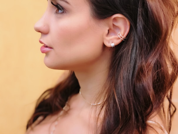 A profile of a person featuring long, dark hair and silver hoop earrings against a soft yellow background. The skin tone is smooth, and the hair slightly waves around the shoulders.