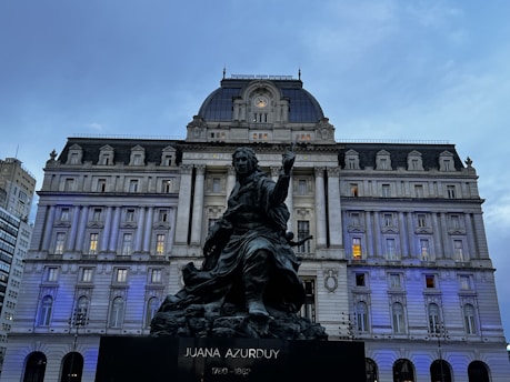 A large historical building with classical architecture featuring columns and intricate detailing stands prominently. In the foreground, a statue of a person, labeled 'Juana Azurduy 1780-1862', is seen. The sky is overcast, and there is a blue tint to the scene, likely due to lighting.