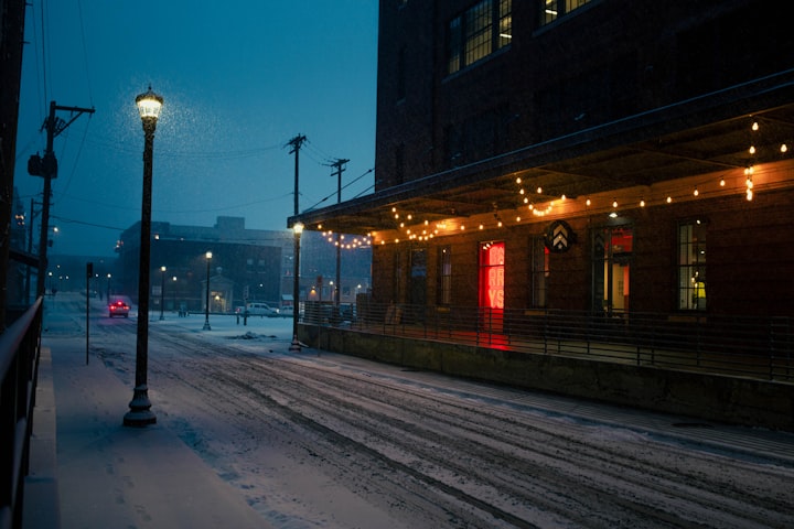 A photo of a snowy street at dusk with a lightly lit building and street lights