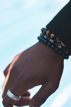 A hand is adorned with multiple beaded bracelets and a silver ring on the finger. The skin tone is deep, and there's subtle detailing in the texture of the hand. The bracelets include various dark-colored beads and tiger's eye stones. The background is softly blurred and light blue.