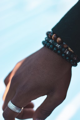 A hand is adorned with multiple beaded bracelets and a silver ring on the finger. The skin tone is deep, and there's subtle detailing in the texture of the hand. The bracelets include various dark-colored beads and tiger's eye stones. The background is softly blurred and light blue.