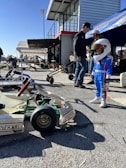At a motorsport pit or garage area, a young child wearing a racing outfit and helmet stands near a go-kart. Several adults stand nearby, some under a canopy. The setting includes a building with a staircase, and multiple people and go-karts are present, creating a busy and lively atmosphere.