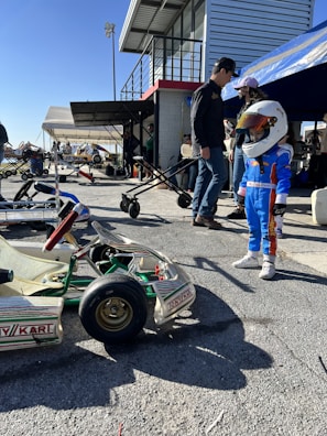 At a motorsport pit or garage area, a young child wearing a racing outfit and helmet stands near a go-kart. Several adults stand nearby, some under a canopy. The setting includes a building with a staircase, and multiple people and go-karts are present, creating a busy and lively atmosphere.