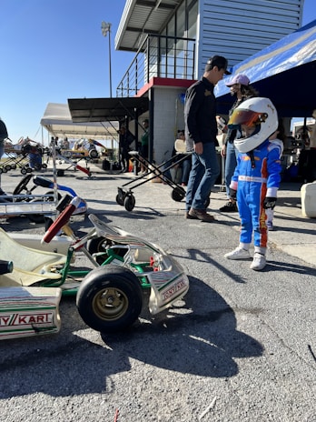 At a motorsport pit or garage area, a young child wearing a racing outfit and helmet stands near a go-kart. Several adults stand nearby, some under a canopy. The setting includes a building with a staircase, and multiple people and go-karts are present, creating a busy and lively atmosphere.