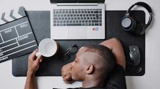 A person rests their head on a desk next to a laptop, a clapperboard, a pair of glasses, a coffee mug, headphones, and a computer mouse. The person appears to be sleeping or resting, with their arm wrapped around the coffee mug. The setting suggests a workspace environment, indicating a break during work or post-work tiredness.