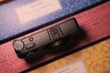 A black camera sits on top of a surface with intricate geometric patterns in blue, pink, and yellow. The camera dial and control buttons are visible, suggesting a focus on photography or travel.