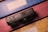 A black camera sits on top of a surface with intricate geometric patterns in blue, pink, and yellow. The camera dial and control buttons are visible, suggesting a focus on photography or travel.