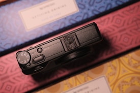 A black camera sits on top of a surface with intricate geometric patterns in blue, pink, and yellow. The camera dial and control buttons are visible, suggesting a focus on photography or travel.