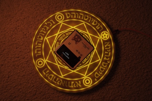 A smartphone is placed on top of a glowing mystical symbol featuring geometric patterns and illuminated text on a circular surface. The floor surface underneath is carpeted, providing a textured background. The overall lighting is dim, emphasizing the glow of the symbol.