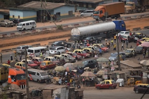 The image depicts a busy street scene with numerous vehicles, including cars and trucks, navigating through a congested area. People are walking around, and there are small roadside stalls with umbrellas. A large tanker truck is prominent on the road alongside various cars, some of which are brightly colored. The environment seems active with a lot of movement and activity.