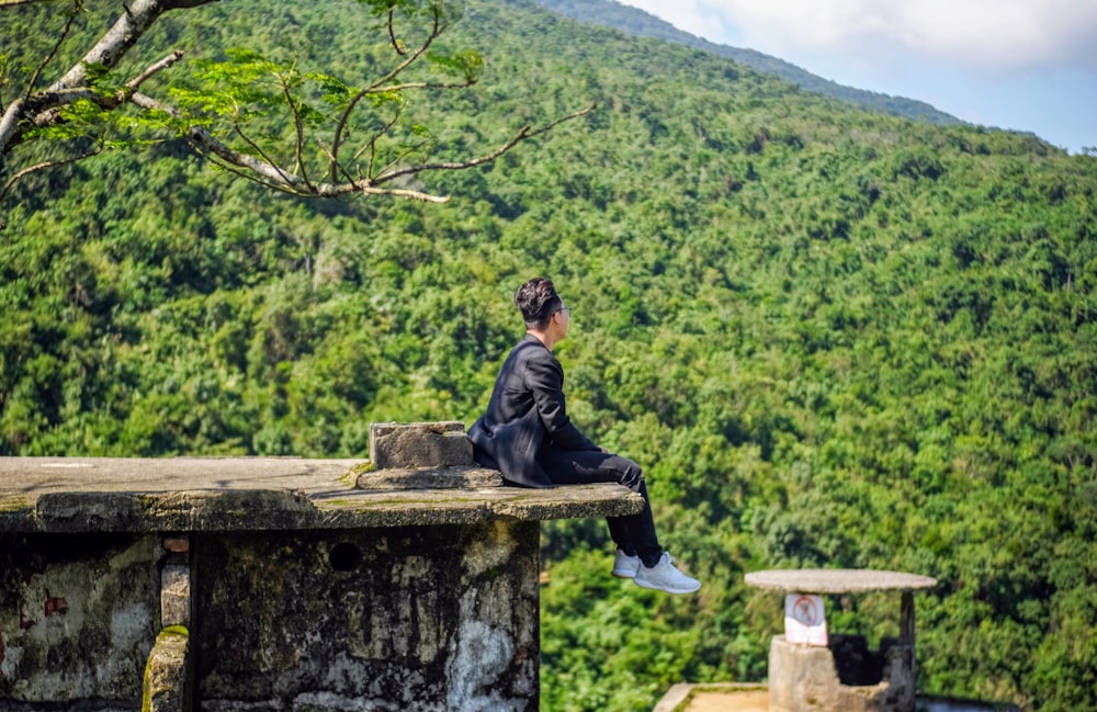 a woman sitting on top of a stone wall next to a lush green hillside