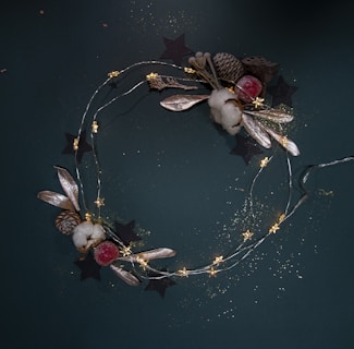 A decorative holiday wreath composed of a thin wire adorned with small, glowing star-shaped lights. It features natural elements such as pine cones, cotton, leaves, and a frosted red berry. The background is a deep green color with subtle sparkles and paper cutout stars.