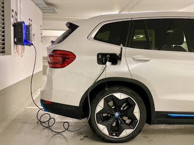 A white electric SUV is parked in a designated parking space within an indoor garage. The vehicle is plugged into a wall-mounted electric vehicle charging station with a coiled black cable. The garage has white walls with black parking numbers visible.
