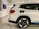 A white electric SUV is parked in a designated parking space within an indoor garage. The vehicle is plugged into a wall-mounted electric vehicle charging station with a coiled black cable. The garage has white walls with black parking numbers visible.