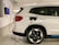 A white electric SUV is parked in a designated parking space within an indoor garage. The vehicle is plugged into a wall-mounted electric vehicle charging station with a coiled black cable. The garage has white walls with black parking numbers visible.