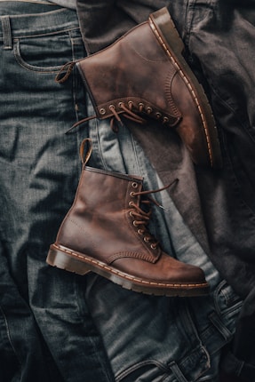 Brown leather boots are set against a background of dark denim jeans, showcasing a contrast in textures. The boots have visible stitching and laces, emphasizing a rugged and vintage style.