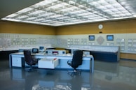 A control room with a large panel of monitors and controls, several office chairs, and desks with electronic equipment. The room is well-lit with a grid of fluorescent lights on the ceiling and has a clean, organized appearance.