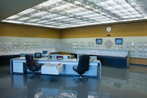 A control room with a large panel of monitors and controls, several office chairs, and desks with electronic equipment. The room is well-lit with a grid of fluorescent lights on the ceiling and has a clean, organized appearance.