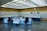 A control room with a large panel of monitors and controls, several office chairs, and desks with electronic equipment. The room is well-lit with a grid of fluorescent lights on the ceiling and has a clean, organized appearance.