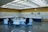 A control room with a large panel of monitors and controls, several office chairs, and desks with electronic equipment. The room is well-lit with a grid of fluorescent lights on the ceiling and has a clean, organized appearance.